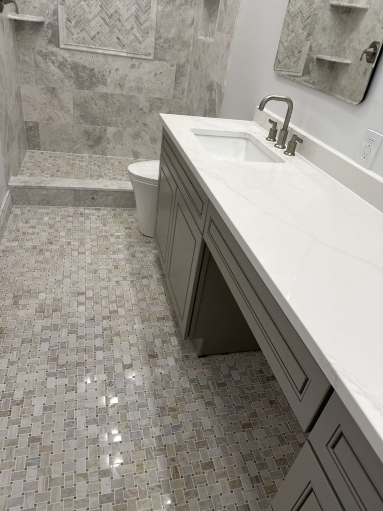 Greige vanity with a white quartz top, a mosaic marble floor, and a walk-in shower beyond.